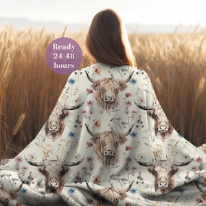 May include: A white throw blanket featuring a repeating pattern of Highland cattle and wildflowers. The cattle are brown and white with long horns. The flowers are in shades of pink, blue, and yellow. The blanket is draped over a person's shoulders. The text "Ready 24-48 hours" is visible in the top left corner of the image.