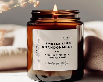 May include: A brown glass candle jar with a white label that reads "Smells Like Abandonment... And I'm Definitely Not Crying! + hand-poured with Love". The candle is lit and the flame is visible.