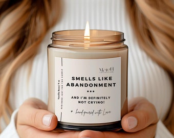 May include: A lit vanilla-scented candle in a clear glass jar with a brown lid. The label reads "Smells Like Abandonment... And I'm Definitely Not Crying!" The candle is hand-poured and held in both hands.