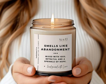 May include: A lit vanilla-scented candle in a clear glass jar. The label reads "Smells Like Abandonment" with the text "Mixed with 100% Betrayal and a Sprinkle of Hope." The candle is hand-poured and held by a person.