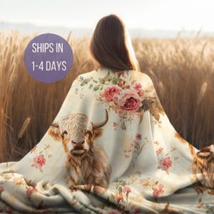 May include: A soft, cream-colored throw blanket with a floral pattern featuring pink roses and a Highland cow. The blanket is draped over a person's shoulders in a field of tall grass.