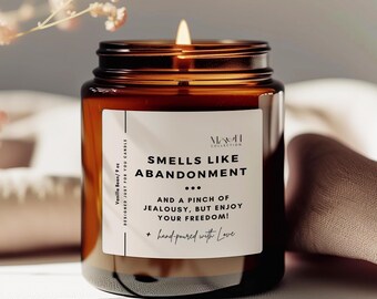 May include: A brown glass candle jar with a white label that reads "Smells Like Abandonment... And a pinch of jealousy, but enjoy your freedom! + hand-poured with love". The candle is lit and the flame is visible.