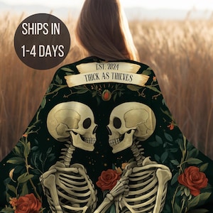 May include: A black throw blanket with a floral design and two skeletons holding hands. The skeletons are facing each other and have a banner above them that says "EST. 2024 THICK AS THIEVES".