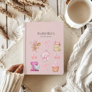 May include: A pink recipe book with the title "Isabella's Recipe Book" in white text. The cover features illustrations of baking supplies, including a whisk, a wooden spoon, a bag of flour, a cherry, a cupcake, and a stand mixer.