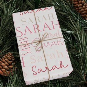 May include: White gift wrap with the name "SARAH" printed in pink and beige script. The paper is tied with a brown twine bow.