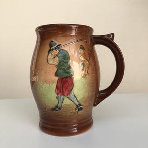 May include: Brown ceramic jug with a handle, depicting a golfer in a hat, green jacket, and red shorts. The background shows a golf course scene. The jug is a decorative item.