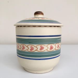May include: Cream ceramic jar with a lid, decorated with blue, green, and pink heart-shaped stripes. The lid has a wooden knob. A decorative kitchen storage container.