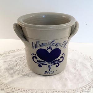 May include: A stoneware crock with two handles, featuring a blue heart design and the text "Williamsburg, VA" and "2000." The crock has a light beige color and sits on a white lace doily.