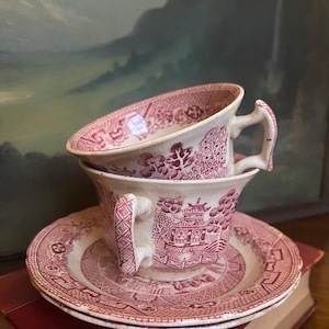 May include: Two stacked antique teacups and saucers with a red and white Asian-inspired design. The cups have handles and are placed on top of each other. The saucers are stacked below. The background is a blurred painting.