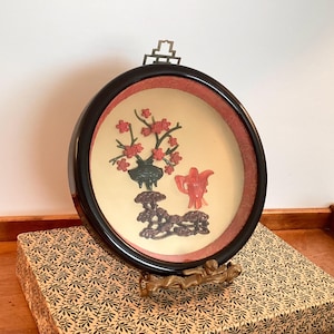May include: A round, framed artwork with a black frame and a cream-colored background. The artwork features a floral arrangement, a small red teapot, and a decorative base. The frame is approximately 10 inches in diameter.