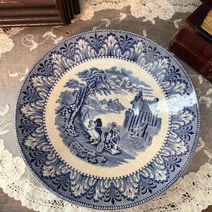 May include: A blue and white antique plate with a detailed landscape scene in the center. The plate has a decorative border and is set on a white lace doily. The plate's design features a horse-drawn carriage.
