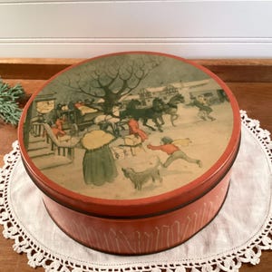 May include: A round, vintage tin with a winter scene illustration on the lid. The tin is red with a decorative pattern around the base. The lid features a detailed depiction of people, horses, and a dog in a snowy landscape.