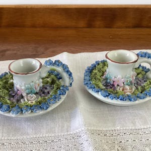 May include: Two white ceramic candle holders with a floral design. Each holder has a small cup with a brown rim, a handle, and is surrounded by green, blue, and pink flowers. The holders sit on a white surface.