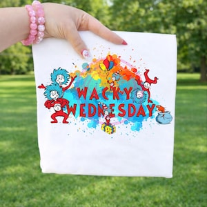 Puede incluir: Tela blanca con un gráfico colorido que presenta personajes y el texto "WACKY WEDNESDAY". El diseño incluye elementos azules, rojos, amarillos y naranjas, con un estilo caprichoso y juguetón. La tela está sujeta por una persona.