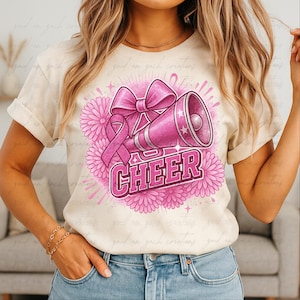 Puede incluir: Camiseta color crema con un diseño rosa de megáfono, lazo y cinta con la palabra "CHEER" en negrita. El diseño está rodeado de flores rosas y destellos de estrellas. La camiseta es usada.