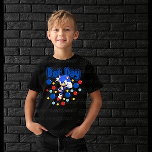 May include: A black short-sleeved t-shirt featuring a graphic of the character Sonic the Hedgehog surrounded by colorful red, blue, and yellow dots. The text Dot Day is printed in blue above the character. The shirt is worn with dark jeans.