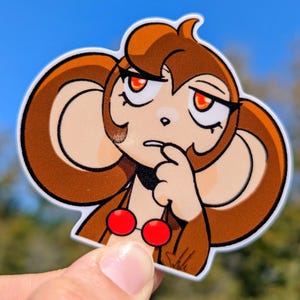 May include: A die-cut sticker featuring a cartoon monkey with brown fur, large ears, and red eyes. The monkey is depicted with a thoughtful expression, touching its finger to its lips. It has two red cherry-shaped objects on its chest.