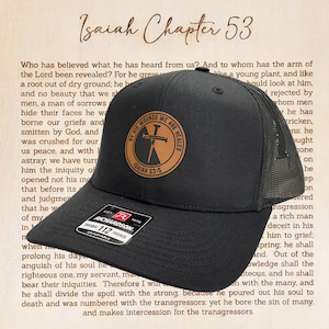 May include: Black trucker hat with a mesh back and a curved brim. The hat features a circular brown leather patch with the text "BY HIS WOUNDS WE ARE HEALED" and "ISAIAH 53:5". The hat is adjustable.