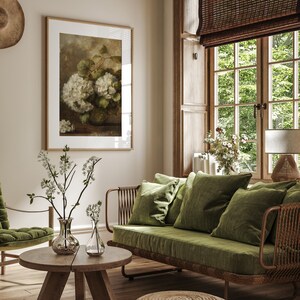 May include: A green velvet sofa with a woven rattan frame and a wooden coffee table with two clear glass vases filled with white flowers. The sofa is in a living room with a large window and a framed print of white flowers.