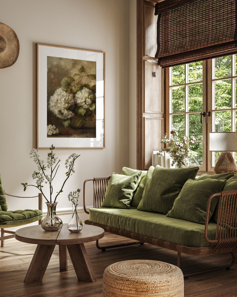 May include: A green velvet sofa with a woven rattan frame and a wooden coffee table with two clear glass vases filled with white flowers. The sofa is in a living room with a large window and a framed print of white flowers.