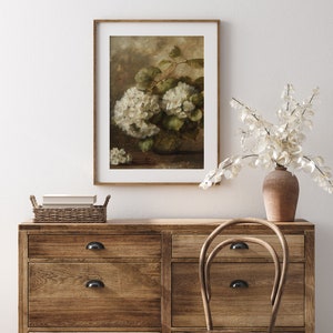 May include: A framed painting depicting a still life with a bunch of white hydrangeas in a brown vase. The painting is set against a light brown background and is displayed on a wooden dresser with a wicker basket and a wooden chair.