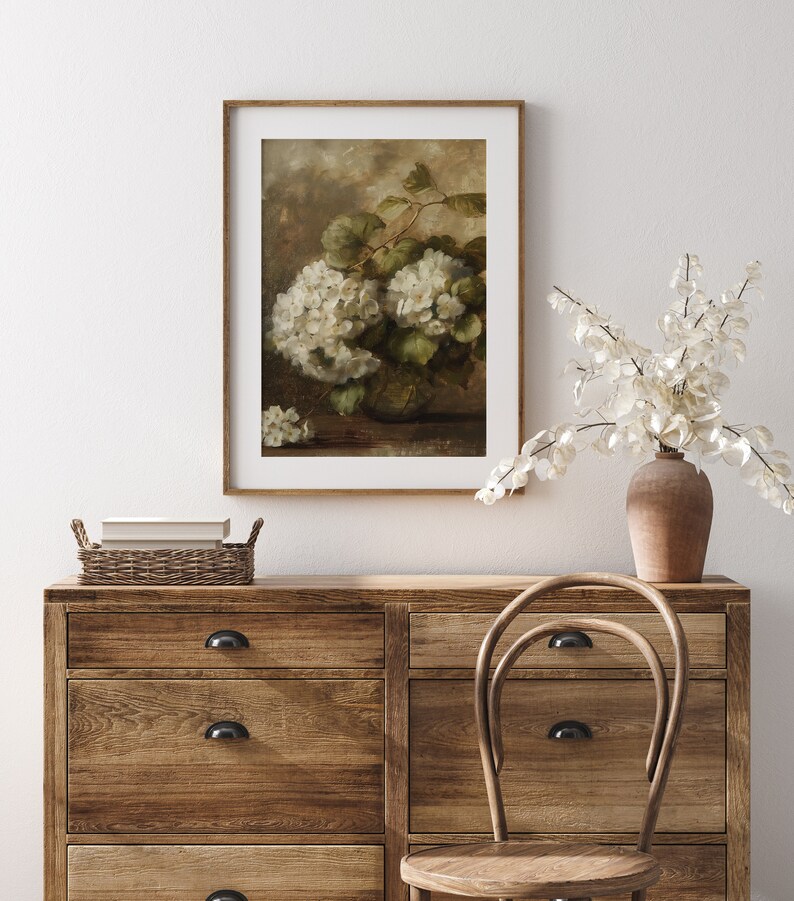 May include: A framed painting depicting a still life with a bunch of white hydrangeas in a brown vase. The painting is set against a light brown background and is displayed on a wooden dresser with a wicker basket and a wooden chair.