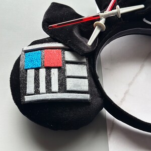 May include: Black headband with a Darth Vader design. The headband features a black bow, a panel with blue and red glitter squares, and a lightsaber. The design is made of soft, plush material.