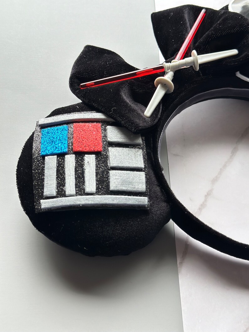 May include: Black headband with a Darth Vader design. The headband features a black bow, a panel with blue and red glitter squares, and a lightsaber. The design is made of soft, plush material.