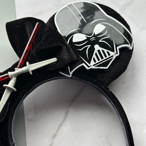 May include: Black headband featuring a Darth Vader design. The headband has a black bow and two toy lightsabers. The Darth Vader image is black and white. The headband is made of a soft, velvety material.