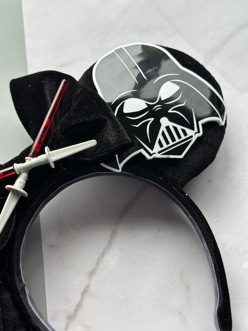 May include: Black headband featuring a Darth Vader design. The headband has a black bow and two toy lightsabers. The Darth Vader image is black and white. The headband is made of a soft, velvety material.