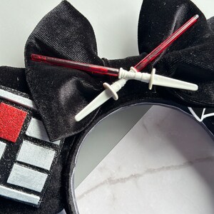 May include: Black headband with a large velvet bow and decorative elements. Two red lightsaber toys are crossed over the bow. The headband features a design with red and silver accents, creating a themed accessory.