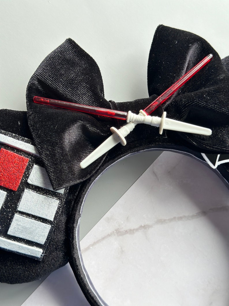 May include: Black headband with a large velvet bow and decorative elements. Two red lightsaber toys are crossed over the bow. The headband features a design with red and silver accents, creating a themed accessory.
