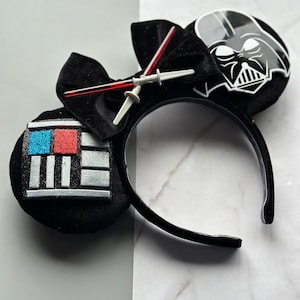 May include: Black headband with two round ears, one featuring a Darth Vader graphic and the other a geometric design with blue, red, and silver accents. A black bow and lightsaber embellishments complete the design.