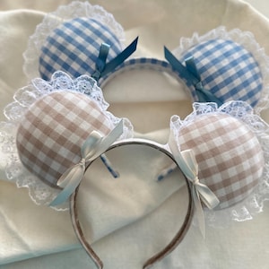May include: Two headbands with plush, gingham-patterned ears. One headband features blue and white ears, the other has tan and white ears. Each ear is trimmed with white lace and a satin bow. The headbands are white and brown.