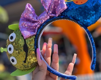 May include: A pair of sequined Minnie Mouse ears with a purple bow, a blue ear with a planet and a green ear with a three-eyed alien face. The ears are held in a hand with long pink nails. The text "@landofjewels" is visible in the image.