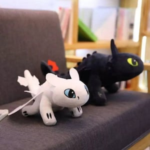 May include: Two plush dragon toys, one black and one white, are sitting on a gray couch. The white dragon has blue eyes and a light gray body. The black dragon has yellow eyes and a dark gray body.