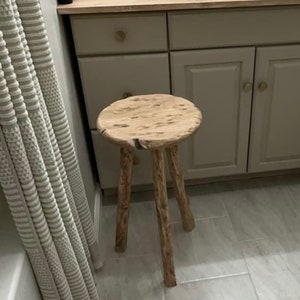 May include: A small, round wooden stool with three sturdy legs. The stool's seat is a light, natural wood color with a unique grain pattern. The legs are also made of wood, with a textured, branch-like appearance. The stool is placed on a tiled floor.