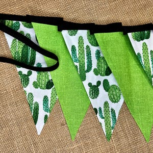 May include: A green and white cactus themed fabric banner with black trim. The banner is made of fabric triangles with a white background and green cactus illustrations. The banner is strung together with black ribbon.