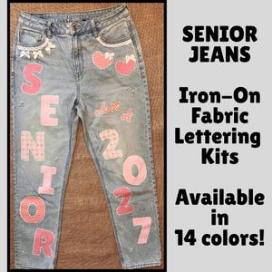 May include: Light blue denim jeans decorated with pink patterned iron-on fabric letters spelling SENIOR and the numbers 2027. The jeans feature white lace trim on the pockets, small white bows, and the text class of. Text reads SENIOR JEANS.
