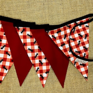 May include: A festive banner with alternating red and gingham patterned pennants. The gingham features a red and white checkered design with black ant illustrations. The banner is strung on a black cord.
