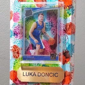 以下が含まれることがあります： ルカ・ドンチッチが青いダラス・マーベリックスのユニフォームを着ている、額縁に入ったバスケットボールカード。カードは、ピンク、ブルー、グリーン、オレンジの斑点のあるカラフルな抽象的な背景で囲まれています。カードには「Rated Rookie」と「Clearly Authentic」のラベルが貼られています。