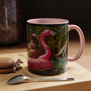 May include: A pink and black coffee mug with a graphic of a hairy man wearing sunglasses and riding a pink flamingo float in a lake.