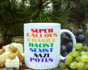 May include: A white ceramic mug featuring the multi-colored text "SUPER CALLOUS FRAGILE RACIST SEXIST NAZI POTUS" printed in a stacked, sans-serif font. The mug has a curved handle on the right side and a smooth, cylindrical body.