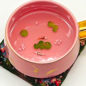 Może przedstawiać: Różowy kubek do herbaty ze złotym uchwytem i wykończeniem, wypełniony różowym płynem i miniaturowymi rybami koi, liśćmi lilii wodnych i kwiatami. Kubek stoi na podkładce w kwiatowy wzór.