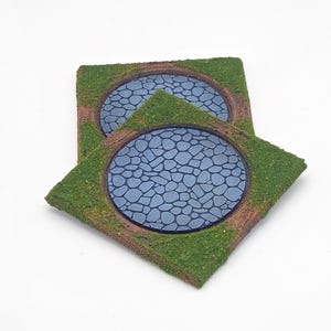 Könnte beinhalten: Zwei Miniatur-Geländebasen, jede mit einem kreisförmigen Teich mit einem Kopfsteinpflastermuster. Die Teiche sind von einem braunen Rand und üppigem grünen Gras umgeben, wodurch eine realistische Miniaturlandschaft für Tabletop-Spiele oder Modellbau entsteht.