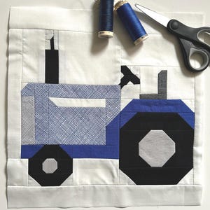 Peut inclure: Un bloc de courtepointe fait à la main représentant un tracteur bleu et noir sur fond blanc. Deux bobines de fil bleu et une paire de ciseaux sont à proximité. Idéal pour un projet artisanal sur le thème de la ferme, mesures en cm.