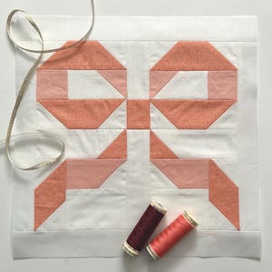 以下が含まれることがあります： ピーチと白の色合いの蝶ネクタイのデザインの手作りのキルトブロック。 2つの糸巻き、1つはバーガンディ、もう1つはコーラルが下部に配置されています。 金色のリボンが上部にかけられています。 キルティングプロジェクトに最適です。