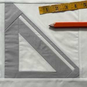 May include: A white fabric square with a gray geometric design. A yellow measuring tape and an orange pencil are on the fabric.