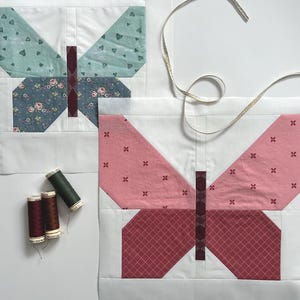Puede incluir: Dos bloques de edredón de mariposa con bordes blancos. Un bloque presenta alas de color turquesa y con estampado floral, el otro alas de color rosa y con estampado rojo. También hay carretes de hilo y una cinta dorada.