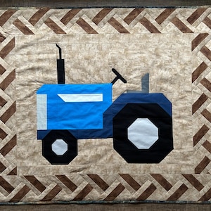 May include: A blue and black tractor quilt with a brown and beige braided border. The tractor is on a beige background.